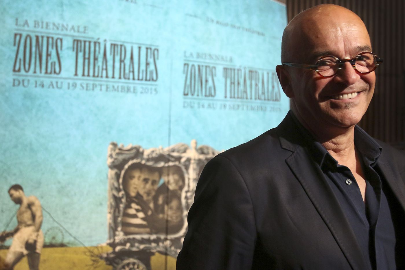 Colour photograph of a bald, smiling, middle-aged man. He wears glasses and a dark suit, standing in front of pale blue posters bearing the words “LA BIENNALE ZONES THÉÂTRALES DU 14 AU 19 SEPTEMBRE 2015”