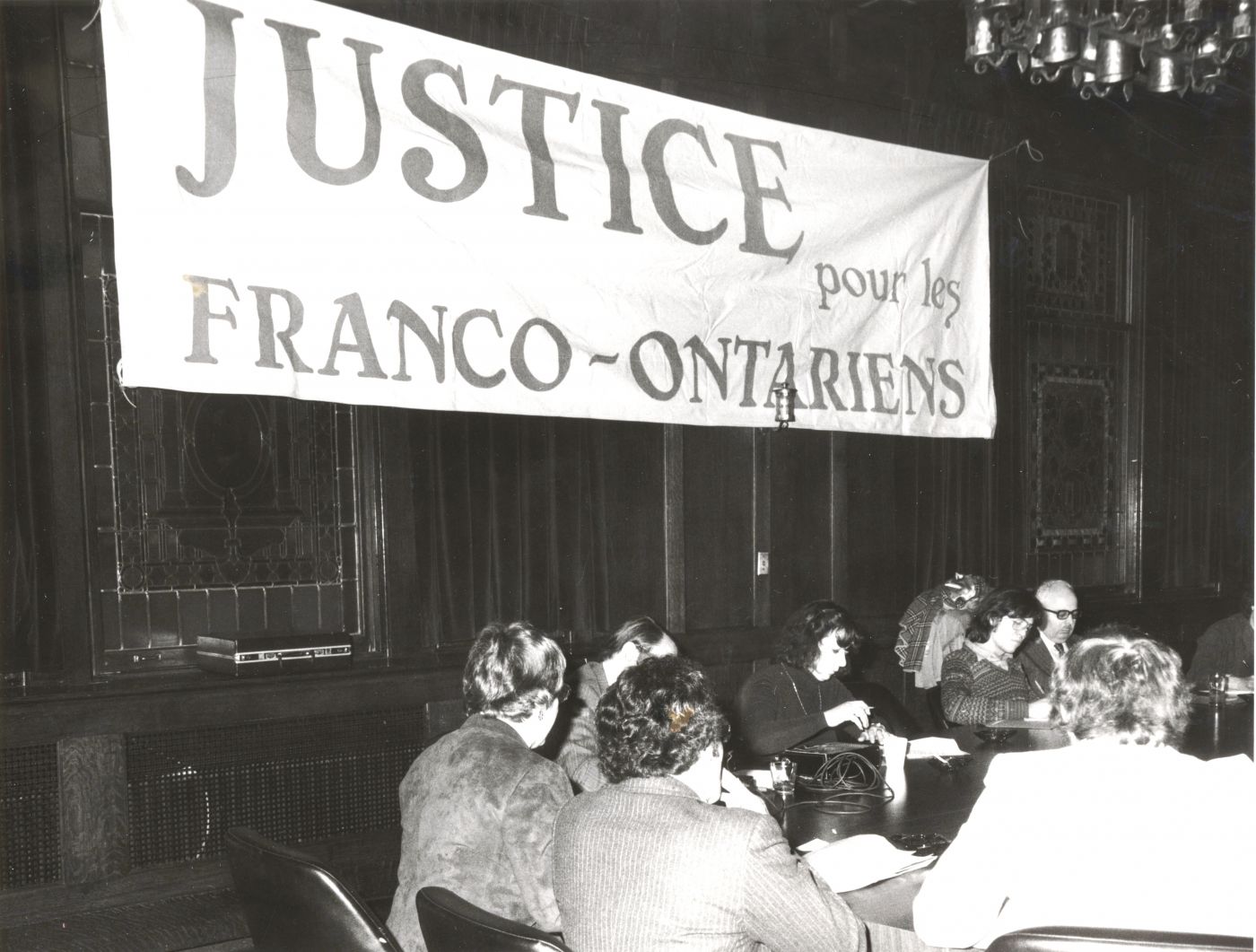 Black and white photograph of a meeting in a room with dark walls covered by a banner with the words “JUSTICE pour les FRANCO-ONTARIENS” (JUSTICE for FRANCO-ONTARIANS). A dozen men and women are seated at a conference table.
