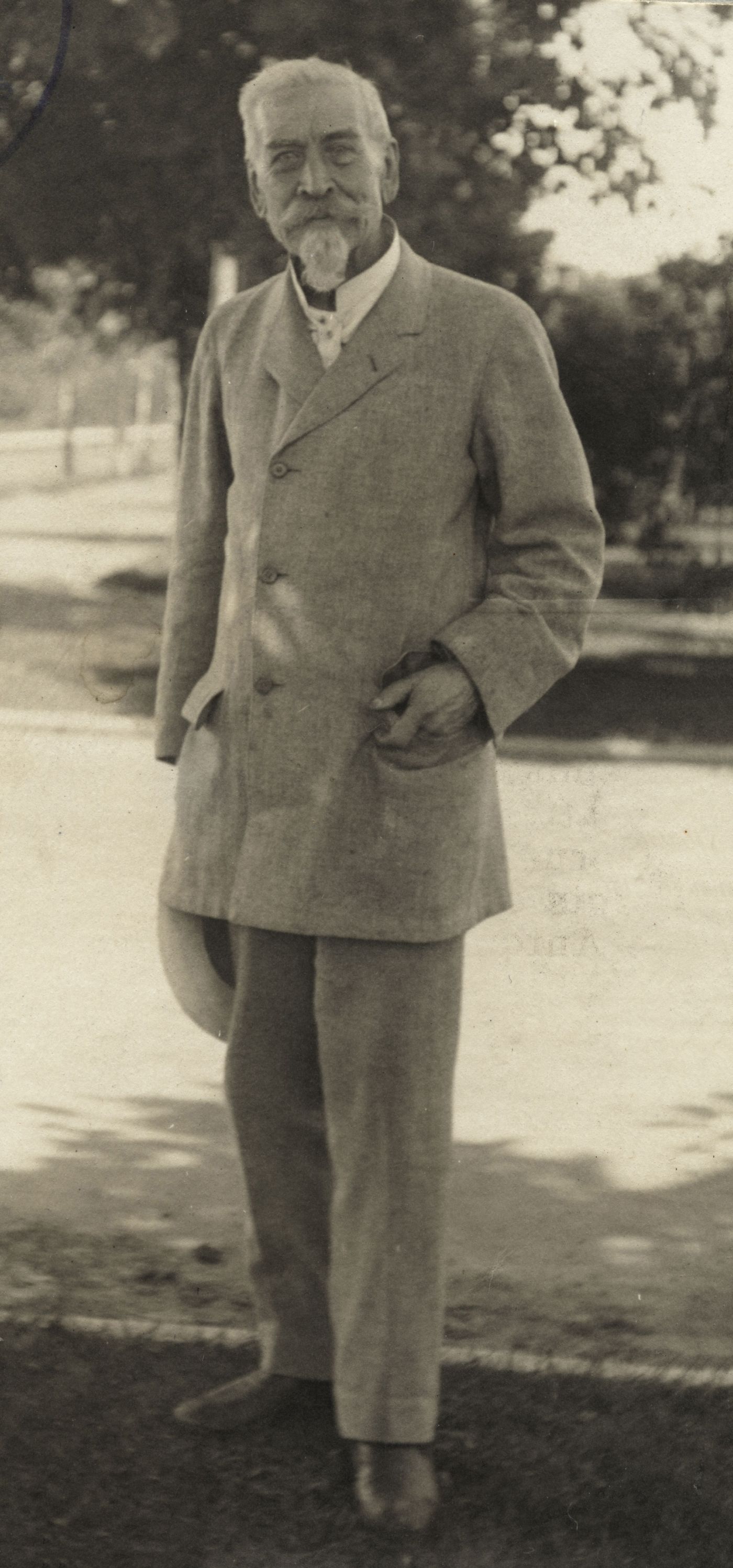 Sepia photograph of an elderly man with white hair, mustache and beard, dressed in a pale suit. He stands at the edge of a tree-lined street, one hand in a pocket, the other holding a hat.