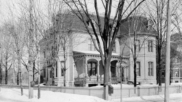 Photographie en noir et blanc d’une maison à trois étages, avec un perron. La maison, vue en hiver, jouit d’une grande cour. Elle est entourée d’une clôture et est bordée d’arbres.