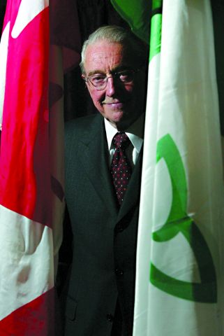 Photographie en couleur d’un homme d’un certain âge souriant. Il porte des lunettes et un costume-cravate. Il est debout entre un drapeau franco-ontarien et un drapeau canadien.