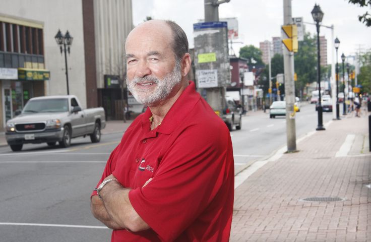 Colour photograph of a middle-aged man dressed in a red polo shirt, arms crossed, with a busy downtown street in the background. He displays an air of determination.