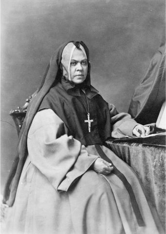 Photographie en noir et blanc d’une religieuse d’âge mûr, assise sur une chaise avec, une table à sa gauche. Elle porte un voile noir ainsi qu’une croix. Elle affiche un air sérieux.