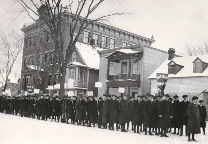 Black and white photograph of a column of fifty young men dressed for the winter. They carry signs and banners with messages such as “Pay our Teachers” and “British Fair Play Please.” In the background, a three-storey brick building and three houses.
