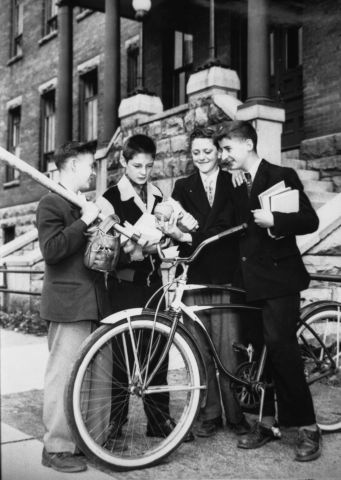 Black and white photograph of four male teens in front of a brick building. Their eyes are turned toward a baseball held  by one of the boys, who is straddling a bicycle. They are dressed in suits and ties. Three of the boys hold books. The fourth holds a baseball bat and glove.