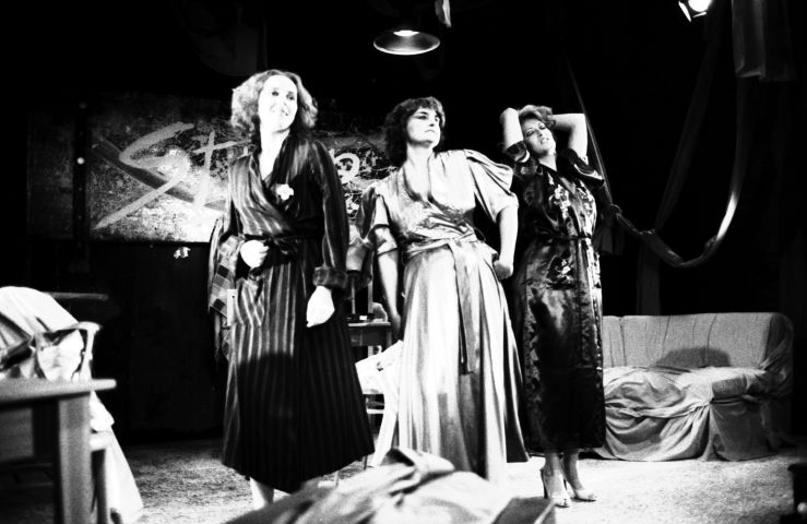 Black and white photograph of three middle-aged women in bathrobes on stage. One of the women has a hand on her hip. Another has her eyes closed and her hands behind her head. The last woman is carrying a towel over her arm. Behind them a sofa; on the wall, a banner.