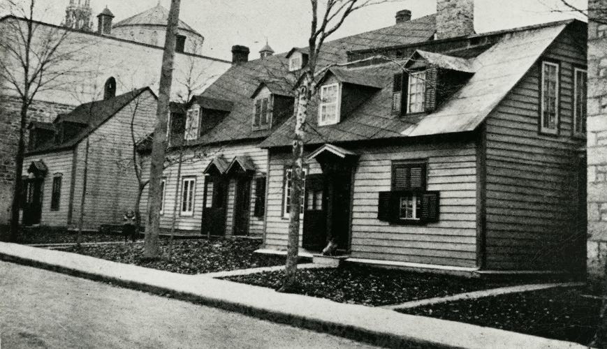 Photographie en noir et blanc de trois petites maisons en bois à deux étages. Les maisons sont très rapprochées les unes des autres, près d’un grand édifice religieux en pierre.