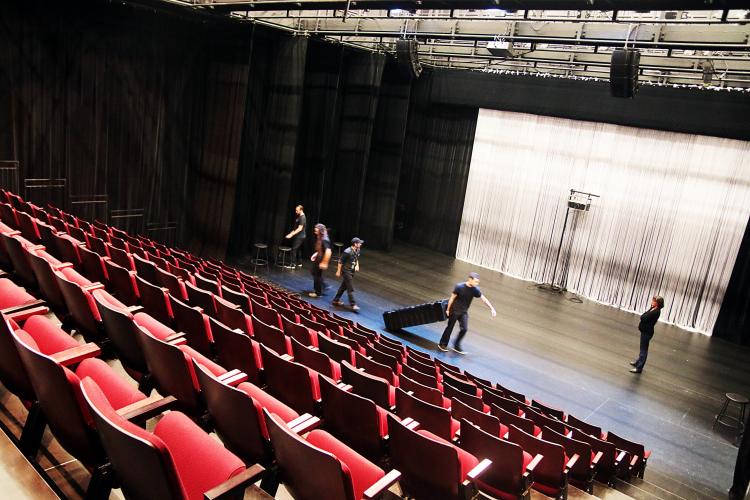 Colour photograph of a theatre, viewed at an angle from above. Rows of bright red seats are arranged in tiers. Five technicians dressed in black work on the stage.