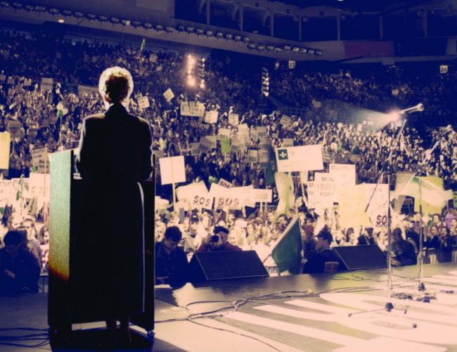 Colour photograph of a woman seen from behind, standing on a stage, addressing the audience from a podium with a microphone. The stadium is filled with people brandishing signs bearing the slogan “S.O.S. Montfort.”