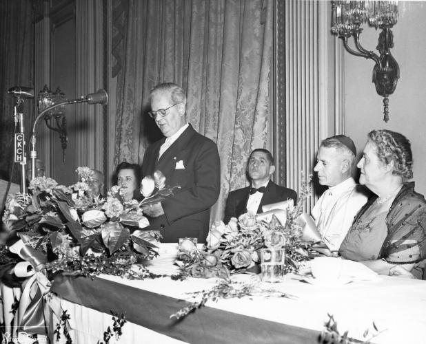 Photographie en noir et blanc d’un homme d’âge mûr en costume et cravate, debout, parlant devant un microphone. Une femme est assise à sa droite, un homme, un religieux portant la calotte et une autre femme sont assis à sa gauche, à une table décorée.