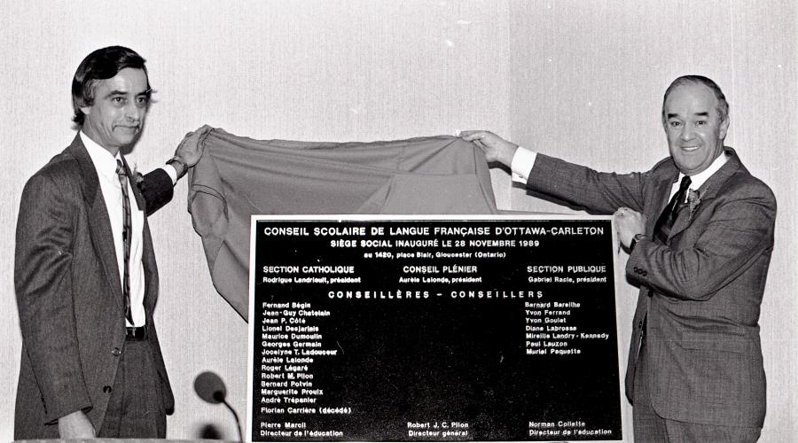 Black and white photograph of two mature men wearing suits and ties. They are smiling as they lift away a cloth to unveil a plaque commemorating the inauguration of the French-language school board. One man looks directly at the camera, while the other looks to one side.